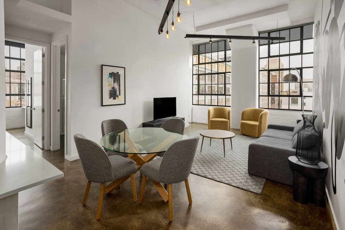 A modern living room with a glass-top dining table, two grey chairs, a sofa, and a TV.
