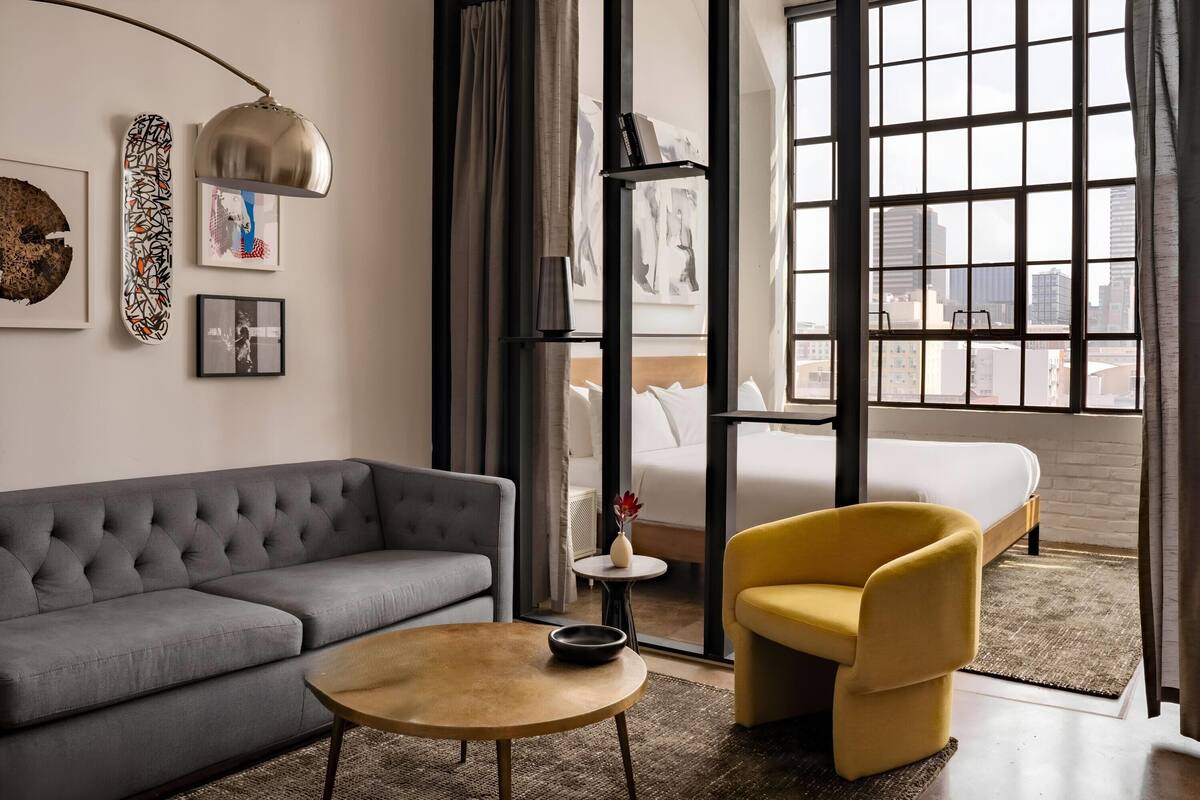 A modern hotel room with a grey sofa, yellow armchair, round coffee table, and large windows offering a city view.