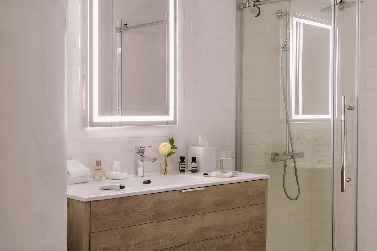 A modern bathroom with a white vanity, wooden drawers, a mirror with integrated lighting, a glass shower enclosure, and a glass door.