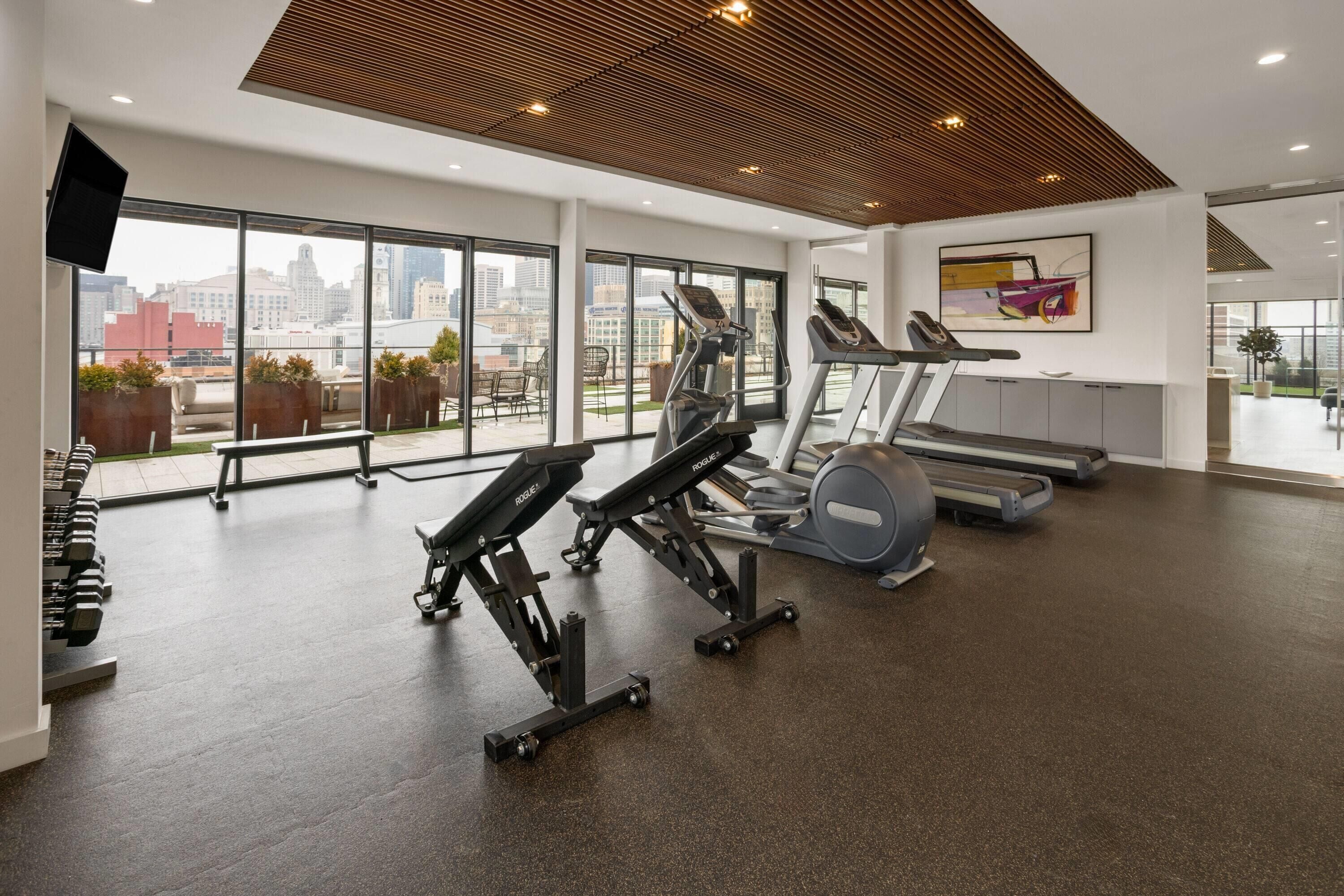 Une salle de sport équipée de tapis de course, d’appareils de musculation et d’haltères, de grandes fenêtres offrant une vue sur la ville et d’un plafond en bois.