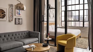 Ett modernt hotellrum med en grå soffa, en gul fåtölj och ett runt soffbord. Det finns en säng med vita sängkläder, ett litet sidobord och ett fönster med utsikt över staden.