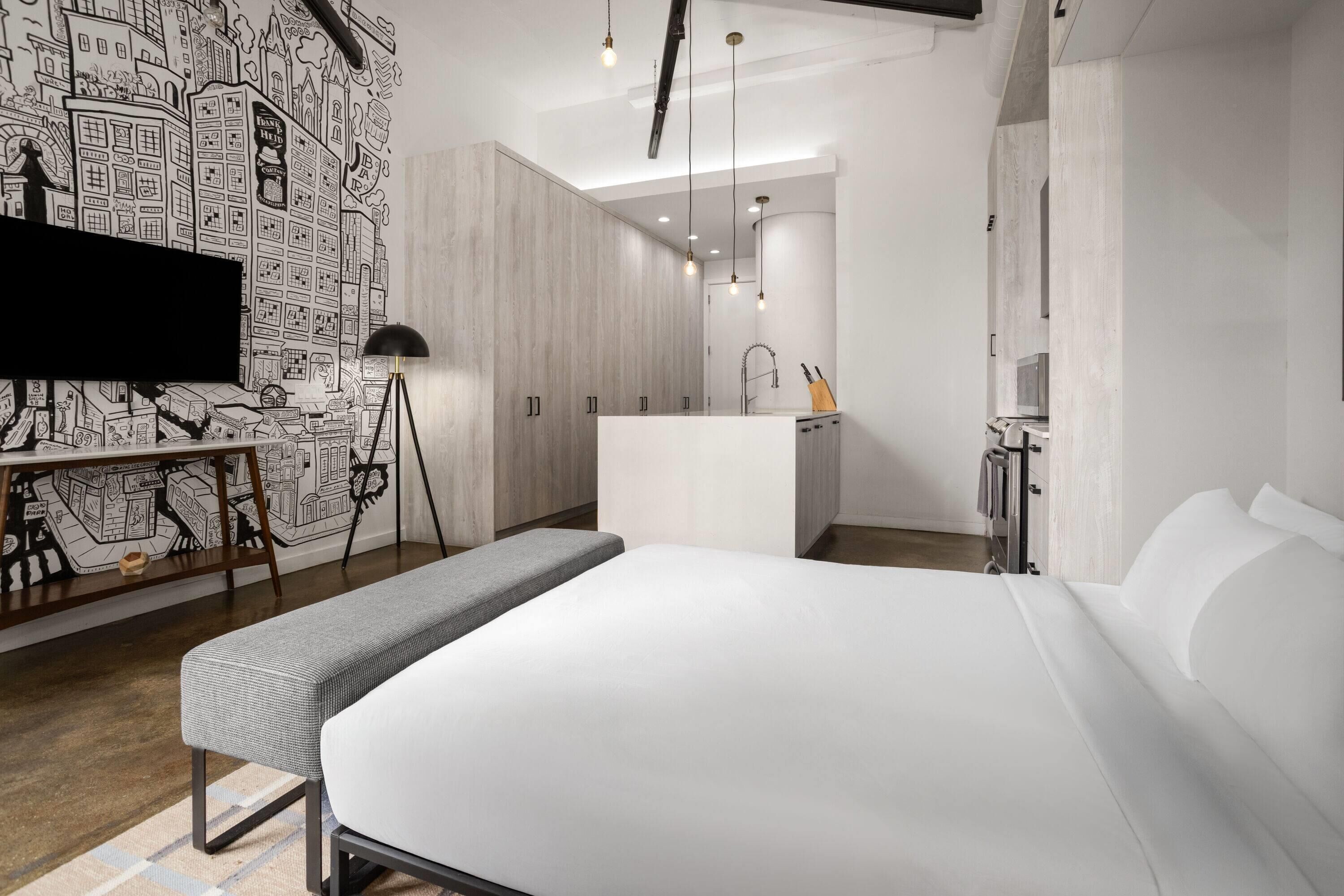 A modern hotel room with a large bed, a desk with a flat-screen TV, a kitchenette, and a city-themed wall mural.