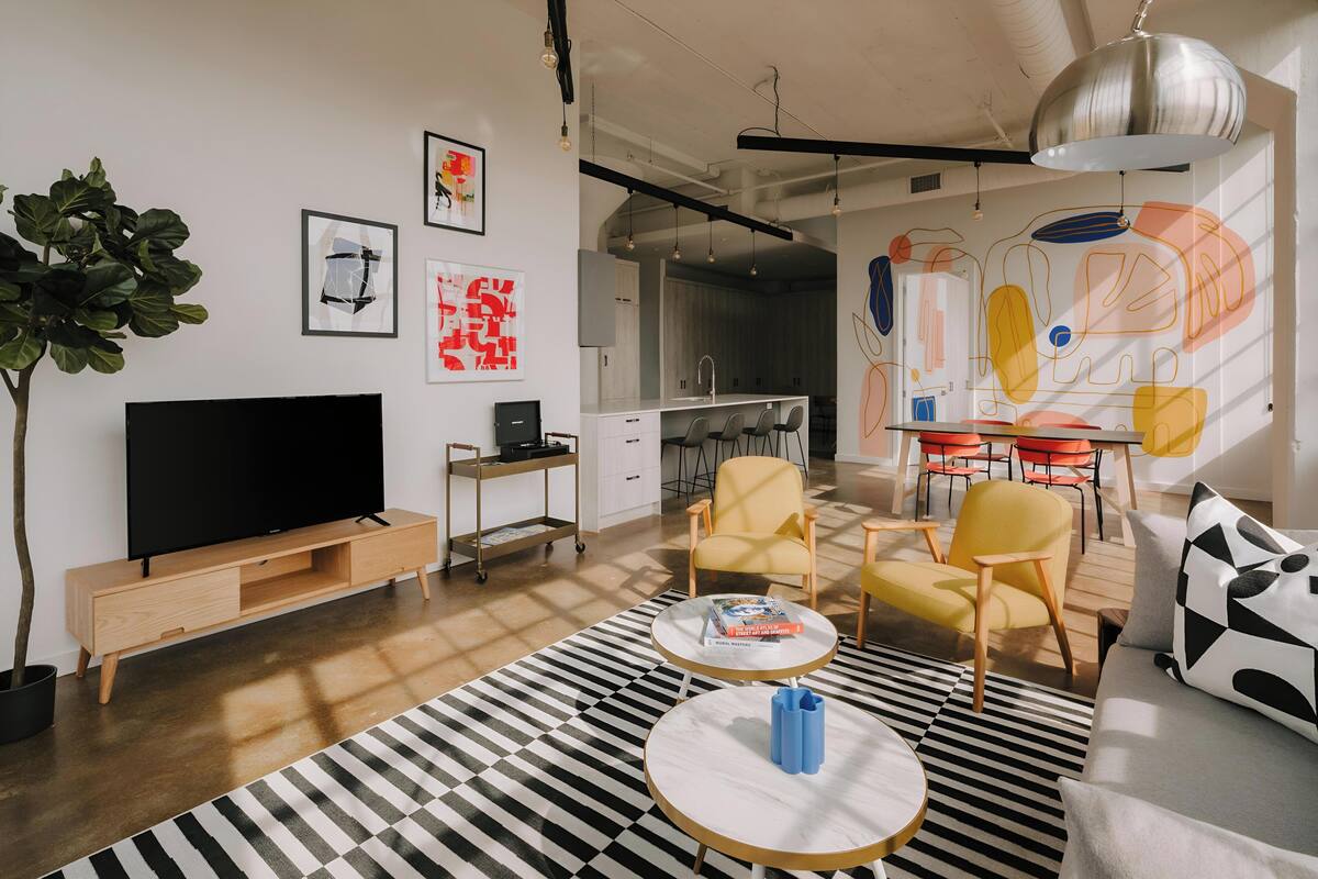 A modern living room with a flat-screen TV, a wooden entertainment unit, a striped rug, and a dining area with yellow chairs and a table.