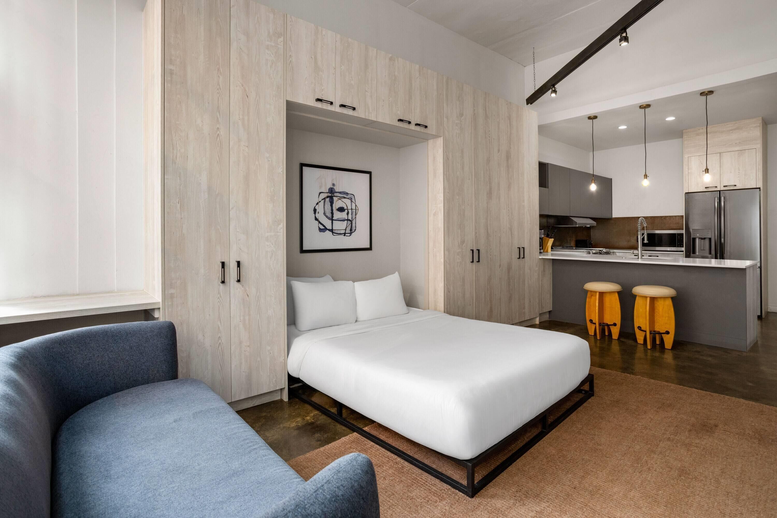 A modern bedroom with a bed, a chair, a desk, and a kitchenette.