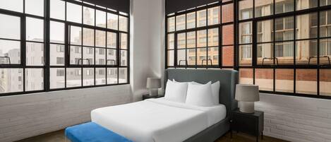 Una habitación de hotel moderna con una cama grande, dos mesitas de noche, un banco azul y ventanas amplias con vista a la ciudad.