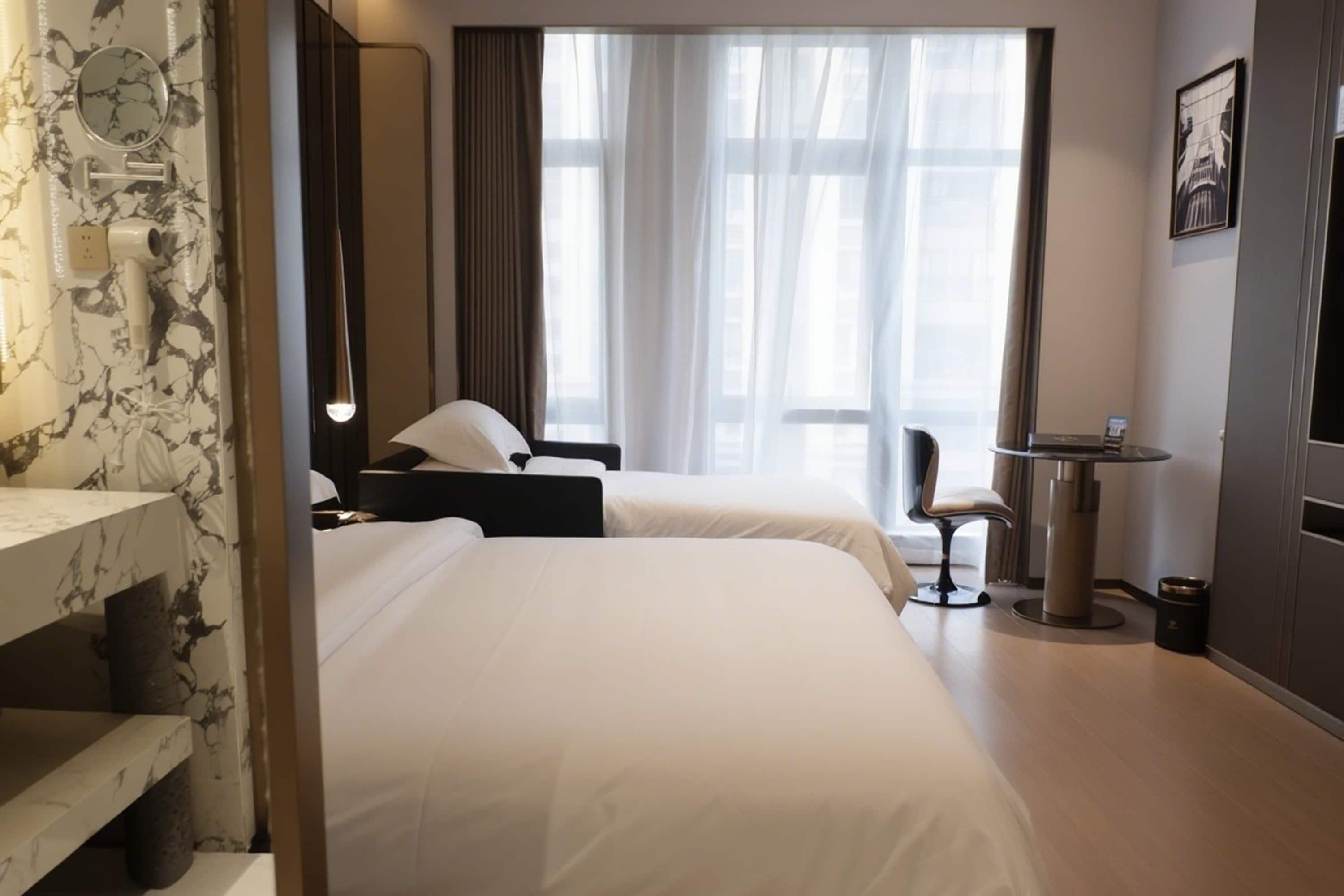 Hotellihuone, jossa on kaksi sänkyä, työpöytä, tuoli, pieni pöytä ja suuri ikkuna, jossa on verhot.