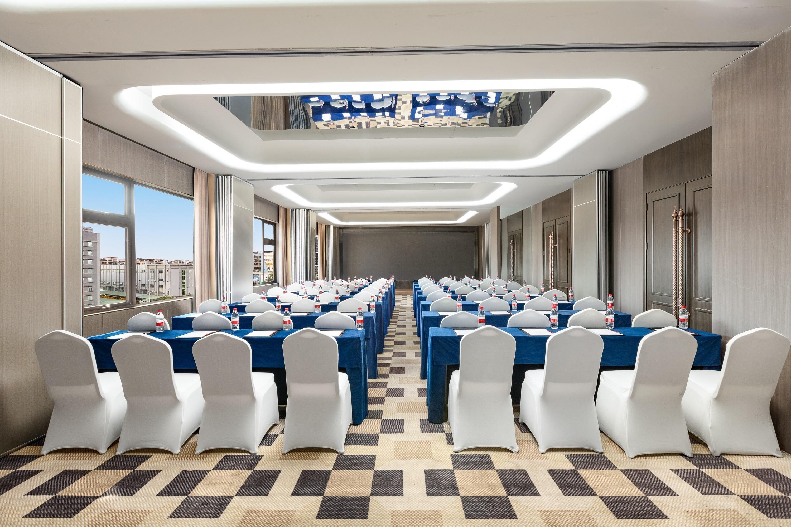 Un salón de conferencias con filas de mesas y sillas, ventanas amplias y una alfombra estampada.