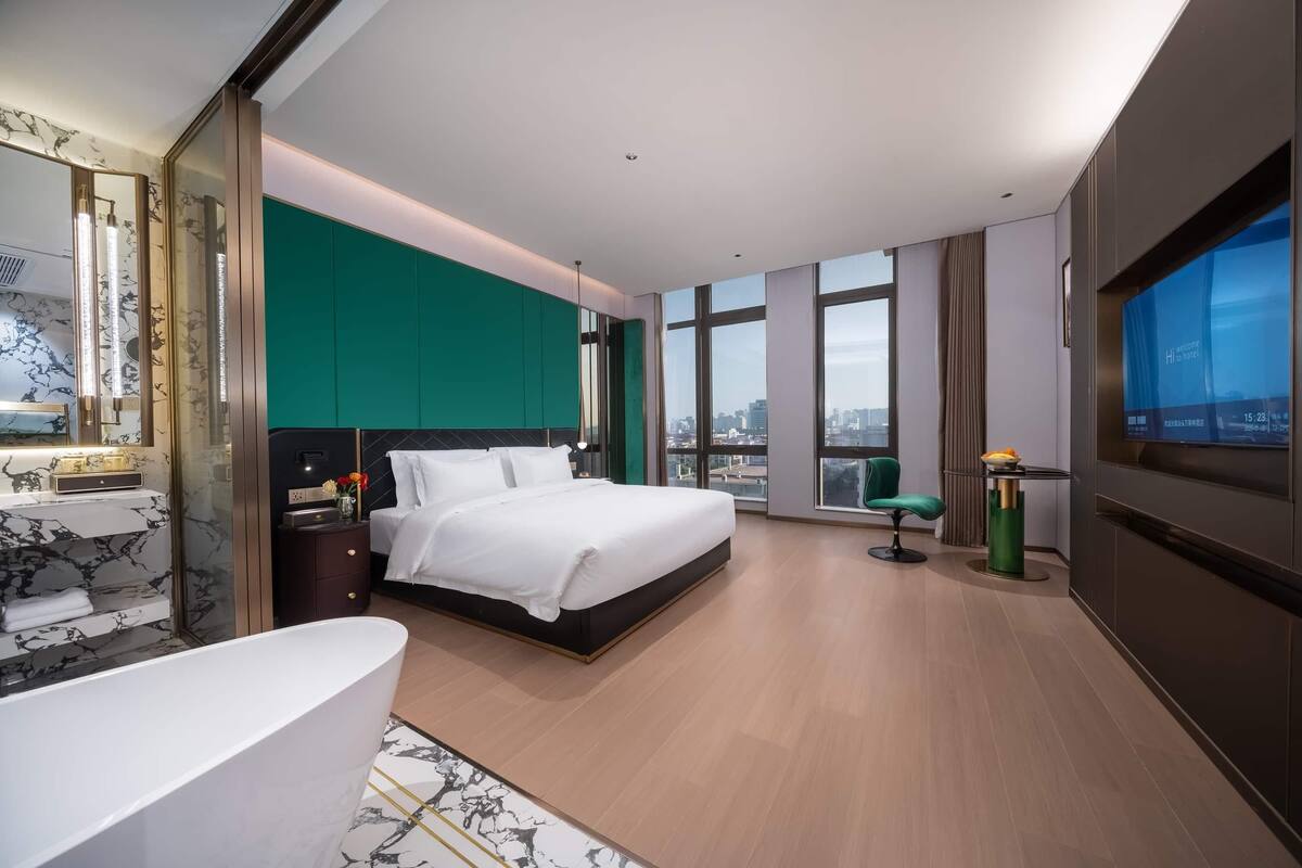 Una habitación de hotel moderna con una cama grande, un área de estar con una silla, televisión y vistas a la ciudad.