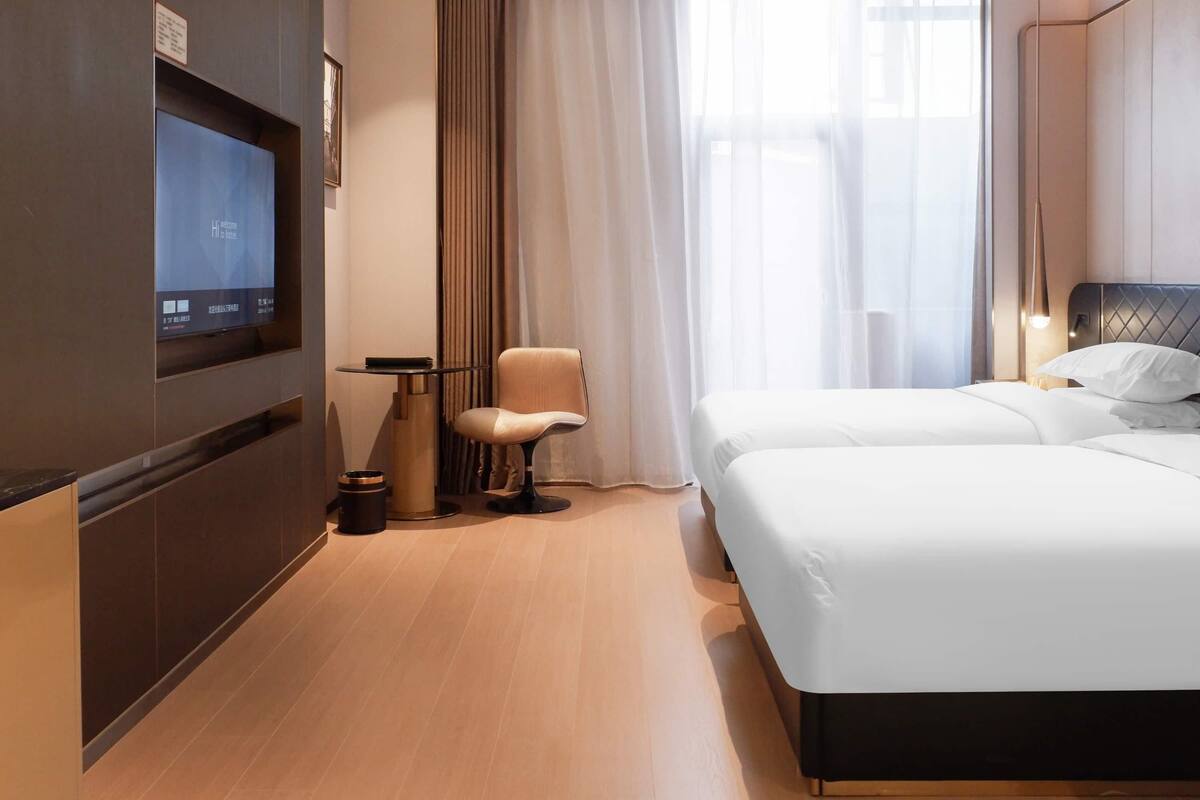 Una habitación de hotel moderna con un televisor de pantalla plana grande montado en la pared, un sillón cómodo, una cama bien hecha y un ventanal con cortinas traslúcidas.