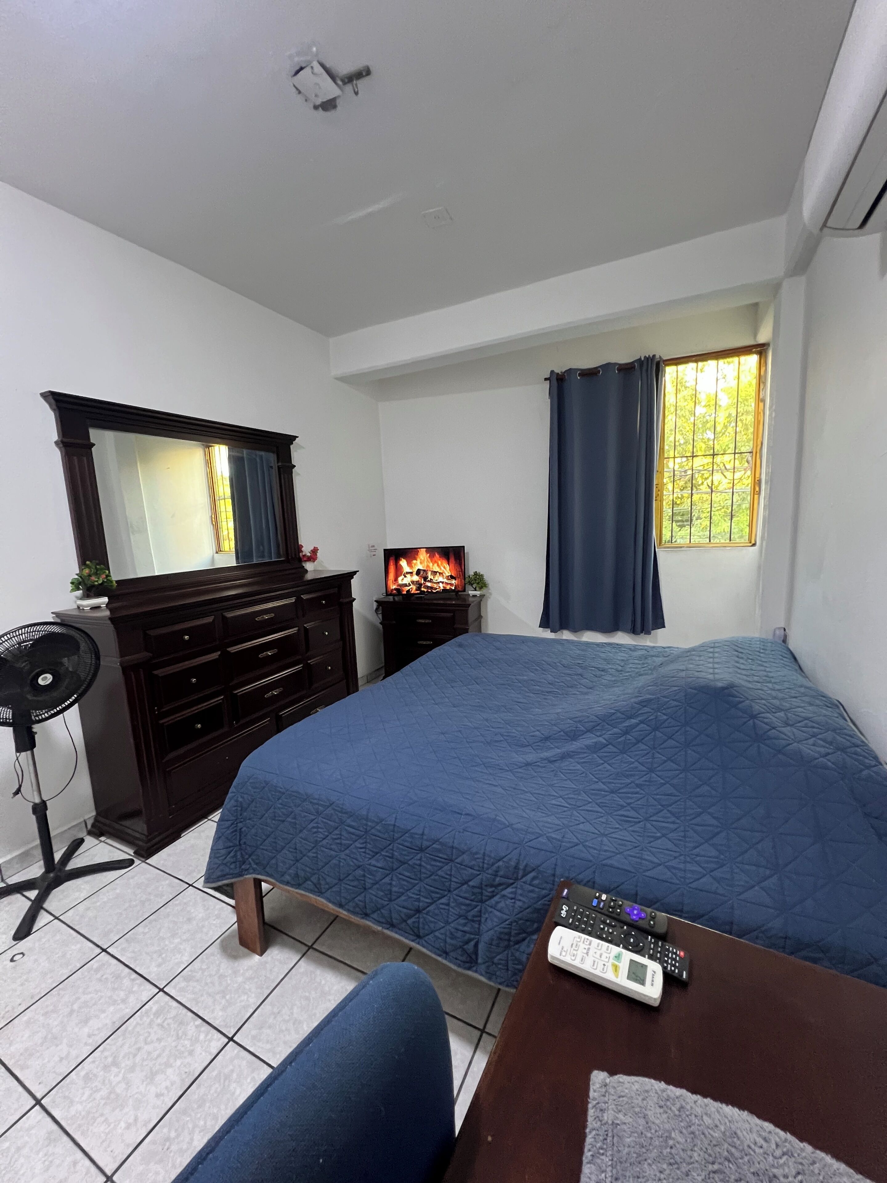 A bedroom with a bed, a dresser, a mirror, a TV, a window with curtains, and a thermostat on the wall.