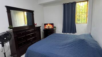 A bedroom with a bed, a dresser, a mirror, a TV, a window with curtains, and a thermostat on the wall.