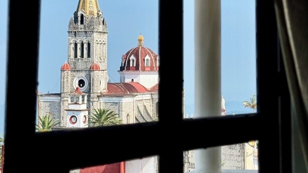 A view through a window of a building with a clock tower and a dome, with a clear sky in the background. - Antica Casa Dáttoli (Cuetzalan del Progreso)