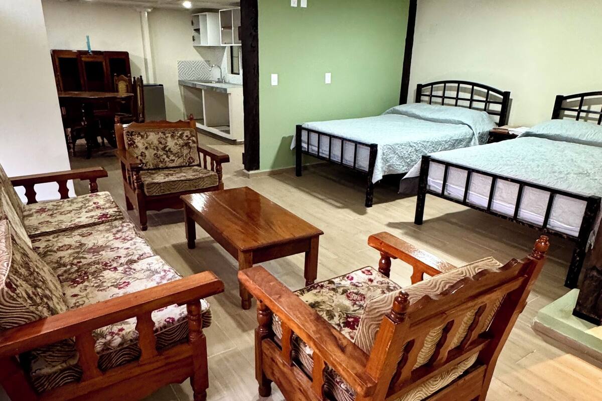 A room with wooden furniture, including sofas and a table, a bed with a blue bedspread, and a kitchenette in the background.