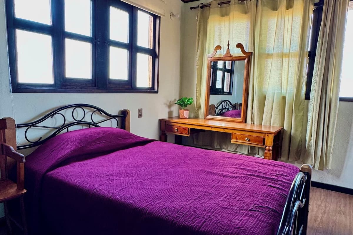 A bedroom with a wooden ceiling, a bed with a purple bedspread, a wooden dresser, a mirror, and a window with curtains.