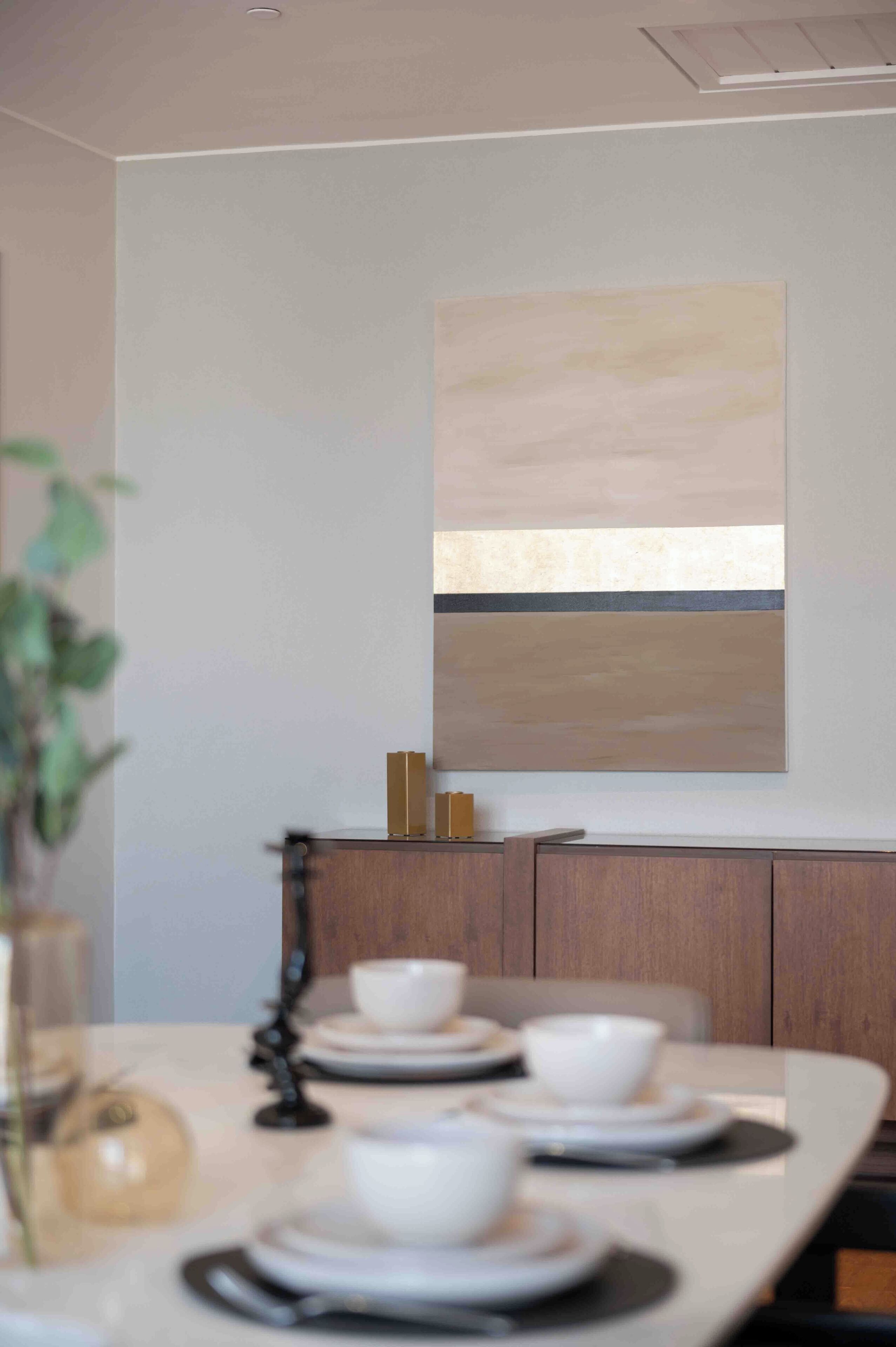 Ein Essbereich mit einem langen Tisch, gedeckt für vier Personen, einem minimalistischen Gemälde an der Wand und einer Pflanze im Vordergrund.