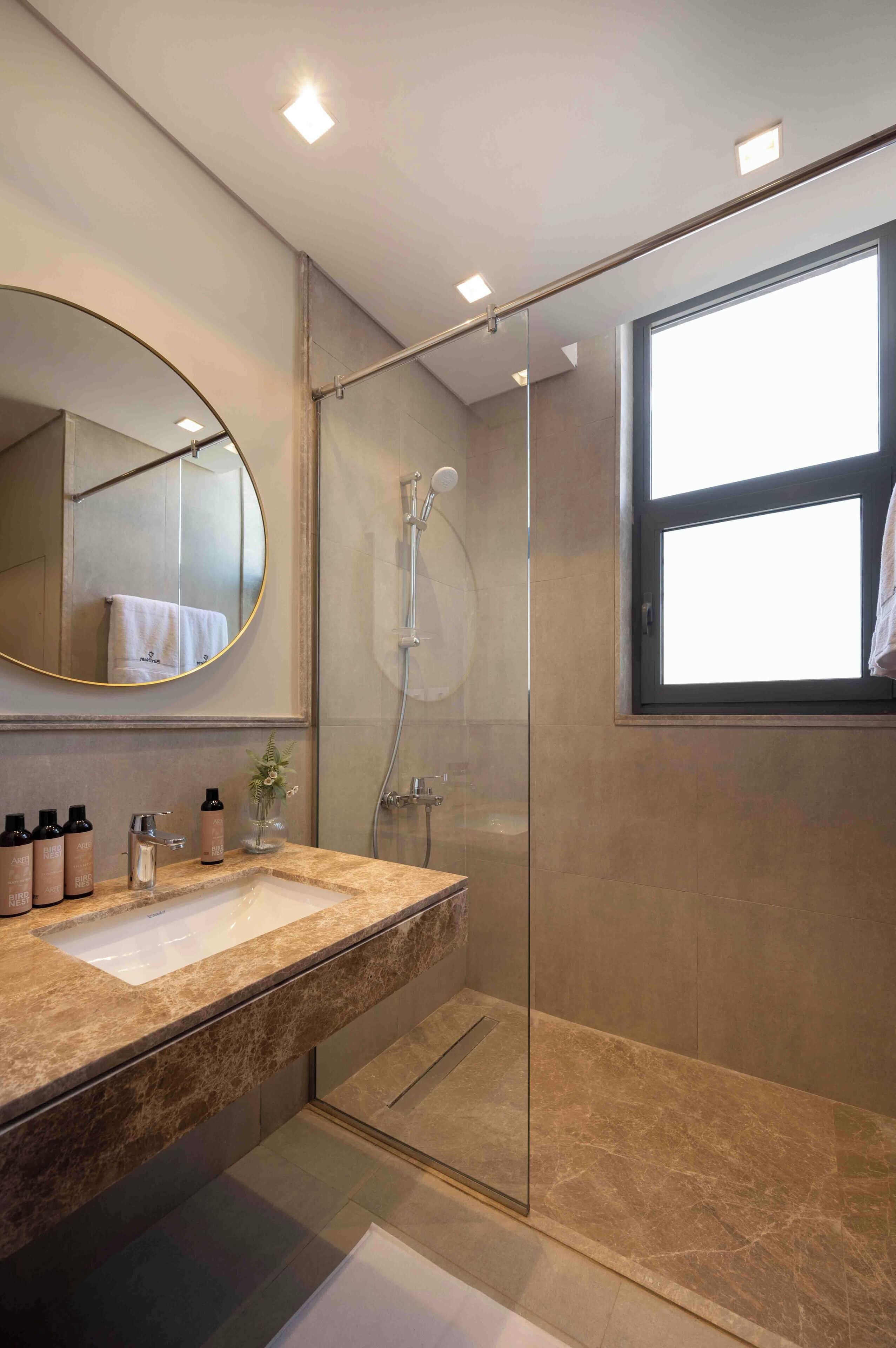 Un baño moderno con ducha de vidrio, un espejo redondo y tocador con encimera de mármol.