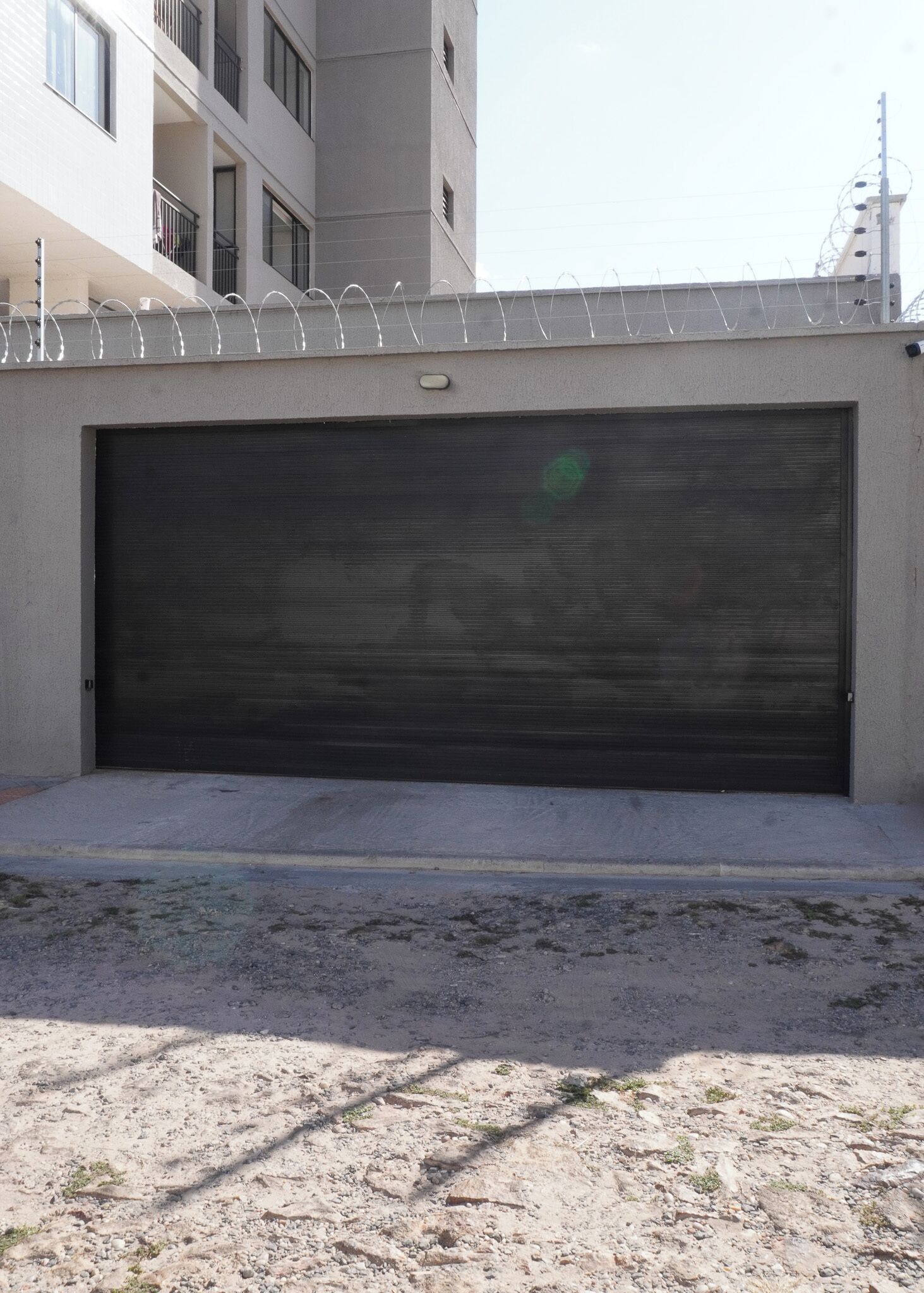 Portão de garagem fechado com barra de segurança na parte superior, piso de concreto e um edifício com várias janelas ao fundo.