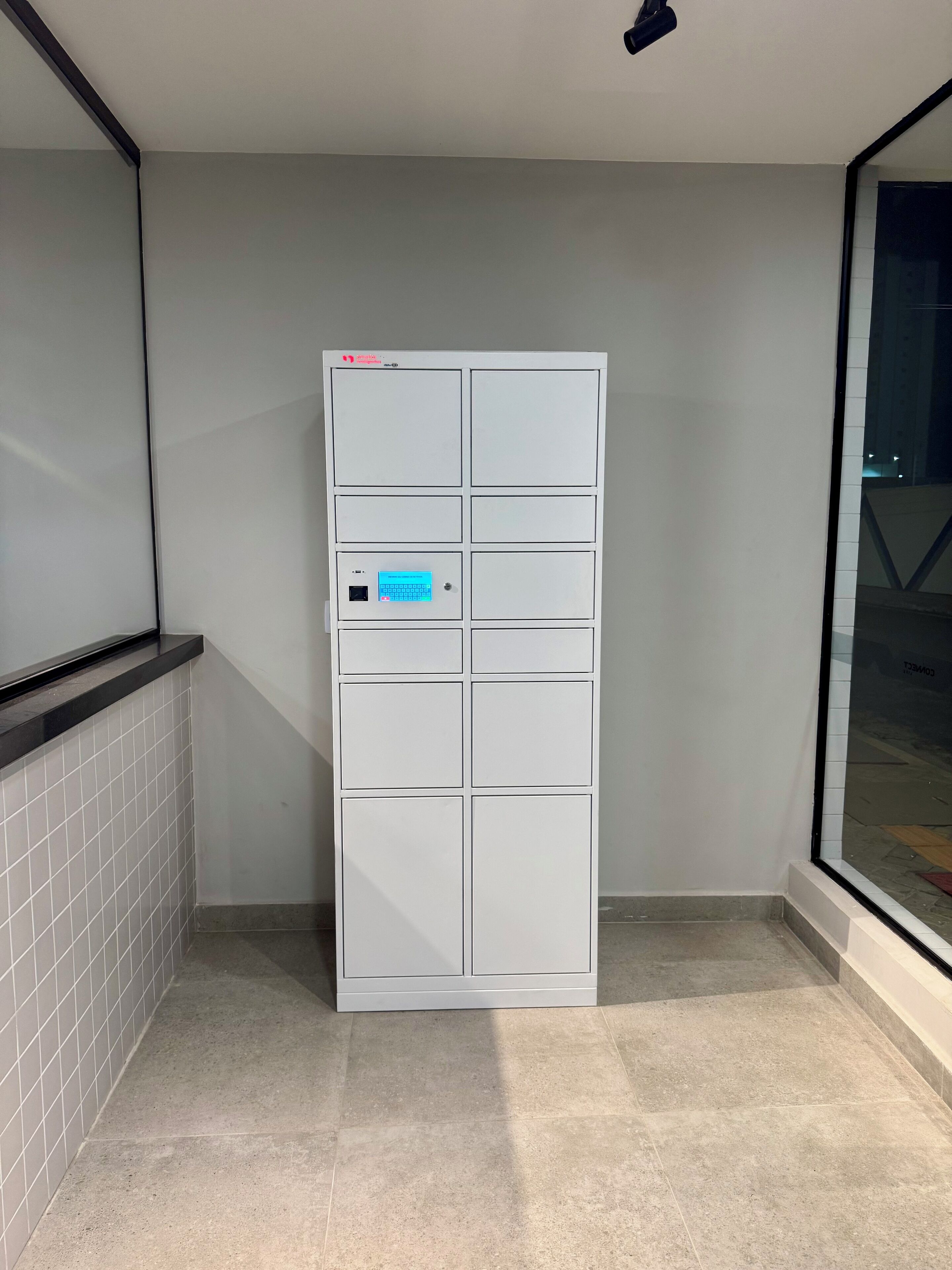 Un mueble de almacenamiento blanco, rectangular y con varios compartimentos, que cuenta con una pantalla digital, situado en una esquina con piso de baldosas y una puerta de cristal que refleja una escena exterior.