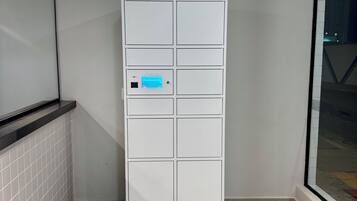 Un mueble de almacenamiento blanco, rectangular y con varios compartimentos, que cuenta con una pantalla digital, situado en una esquina con piso de baldosas y una puerta de cristal que refleja una escena exterior.