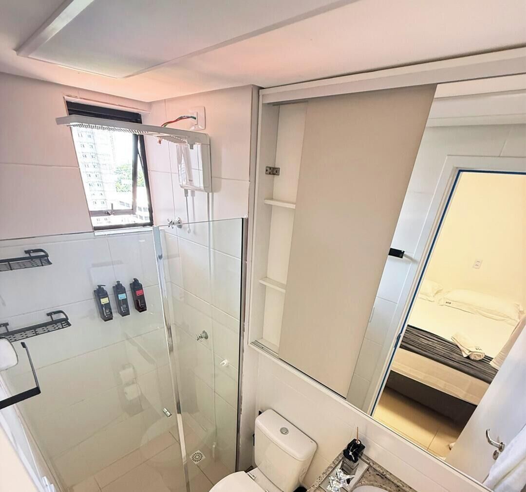 A compact bathroom with a glass shower enclosure, a toilet, and a large mirror reflecting a bed.