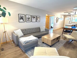 A modern living room with a grey sofa, a wooden coffee table, a beige armchair, and a dining area with a wooden table and chairs. - Cozy Family 2-Bedroom Getaway in Canmore,  perfect for families! (Canmore)
