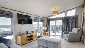A modern living room with a sofa, ottoman, wooden TV stand, and a large window with curtains. - 1 Fishermans Hill (Newton Stewart)
