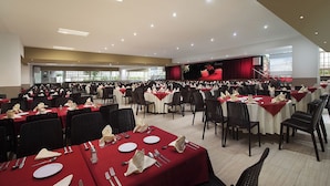 A large banquet hall with rows of tables set for dining, a stage with a red curtain, and a projector screen. - El Cid Castilla Beach Hotel - All Inclusive (Mazatlán)
