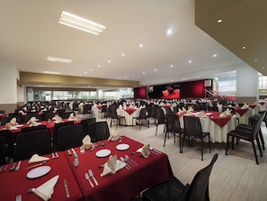 Un amplio salón de banquetes con filas de mesas dispuestas para comer, un escenario con cortina roja y una pantalla de proyector.