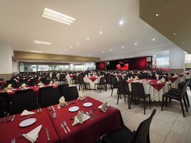 Un amplio salón de banquetes con filas de mesas dispuestas para comer, un escenario con cortina roja y una pantalla de proyector.