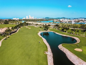 Un campo de golf con un estanque, palmeras y un paisaje urbano al fondo.