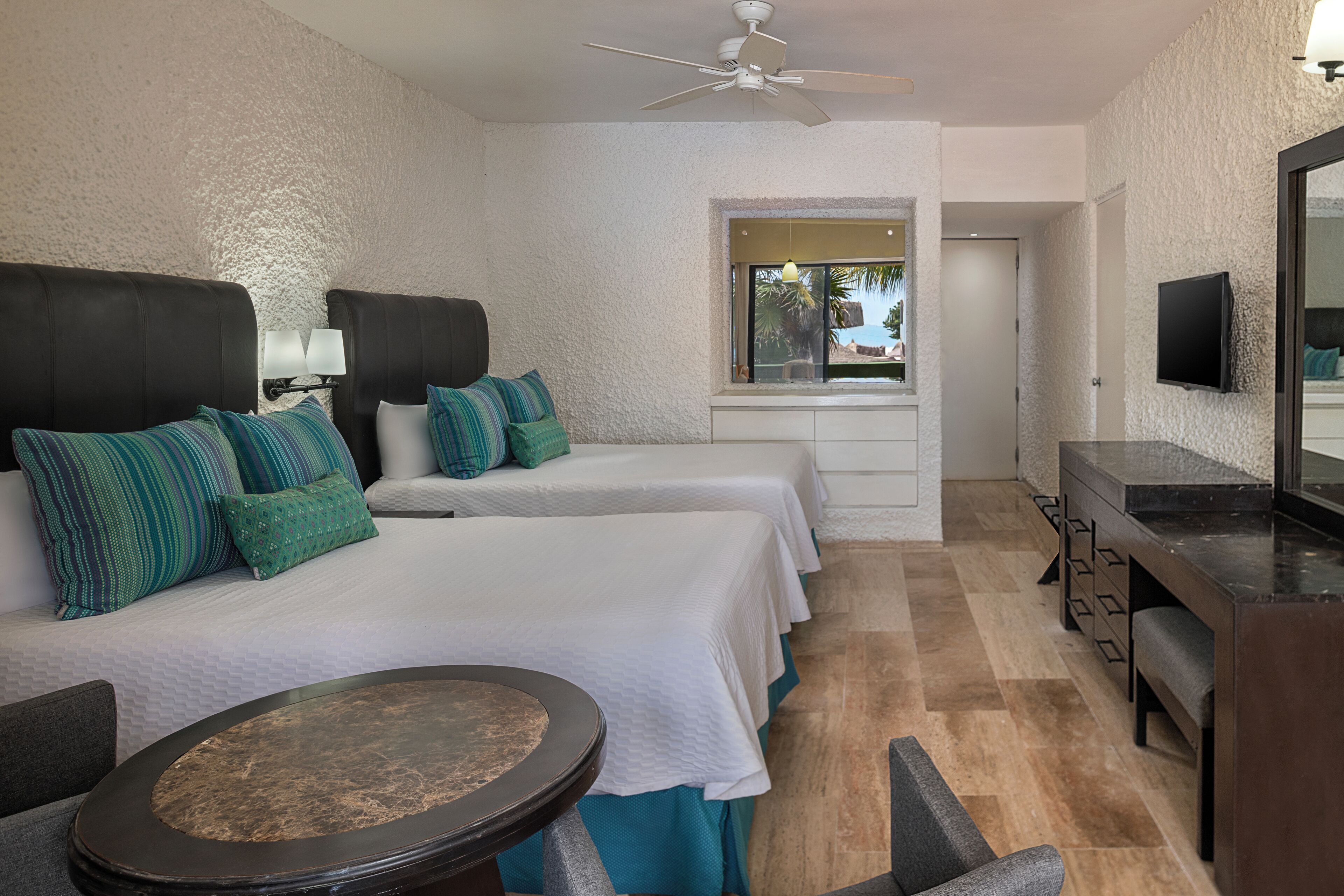 Quarto de hotel com duas camas, uma escrivaninha, uma cadeira, uma mesa redonda, um ventilador de teto e uma televisão fixada na parede.