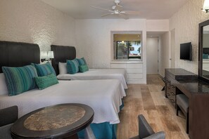 Habitación de hotel con dos camas, un escritorio, una silla, una mesa redonda, un ventilador de techo y un televisor montado en la pared.