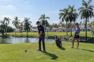 Un campo de golf con un estanque, palmeras y un club de campo al fondo.