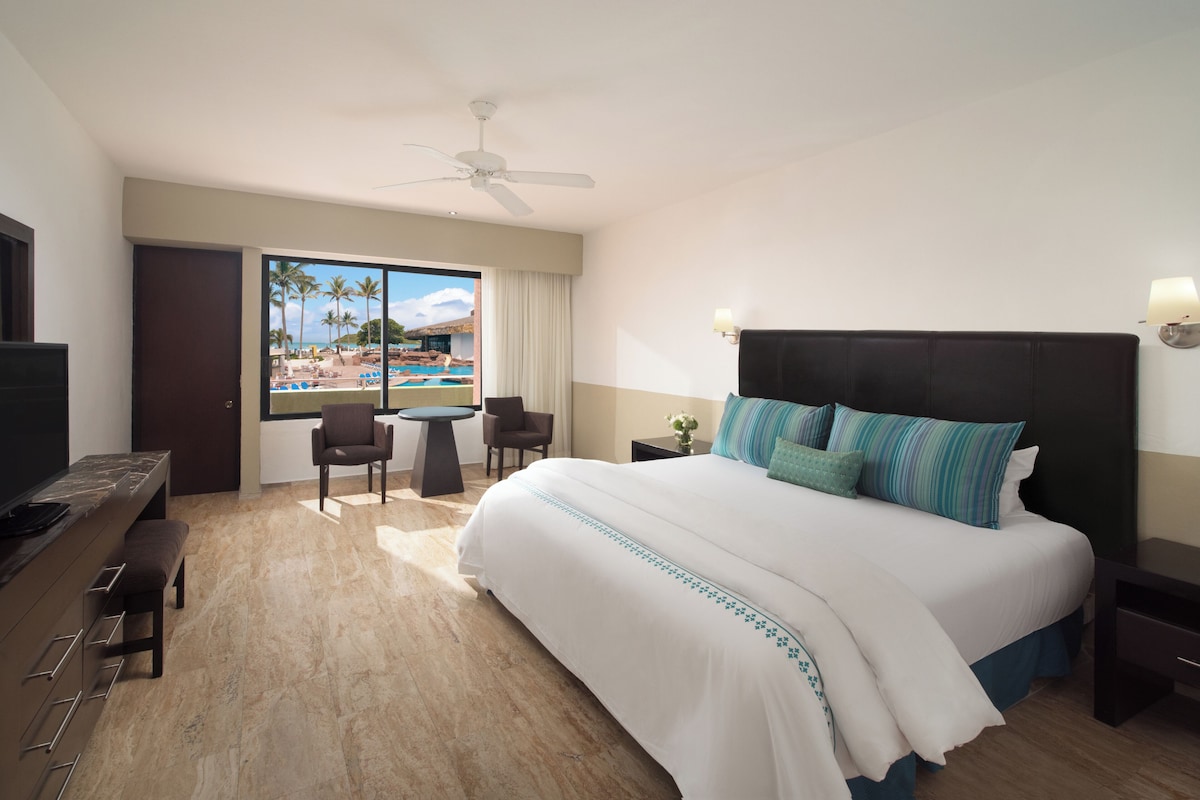 A hotel room with a bed, a desk, a chair, a TV, a ceiling fan, and a window with a view of a pool and palm trees.