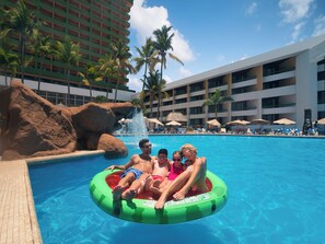 Piscina con una formación rocosa y palmeras, rodeada por un edificio de hotel con varios balcones.