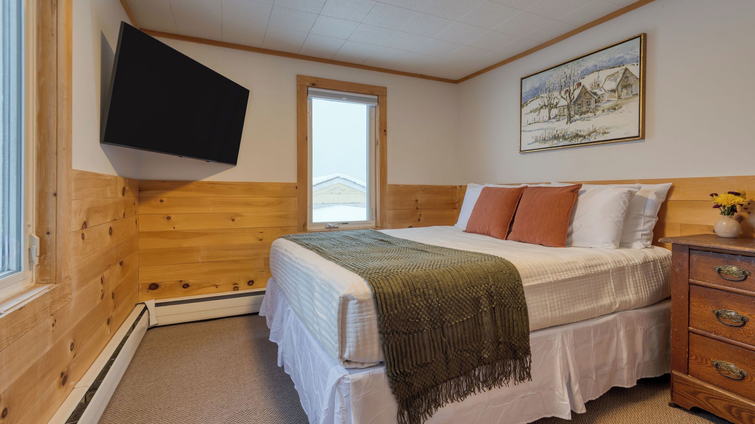 Une chambre avec une tête de lit en bois, un lit avec une literie blanche, un plaid marron, une commode en bois avec des tiroirs, un tableau au mur et une fenêtre donnant sur une maison.