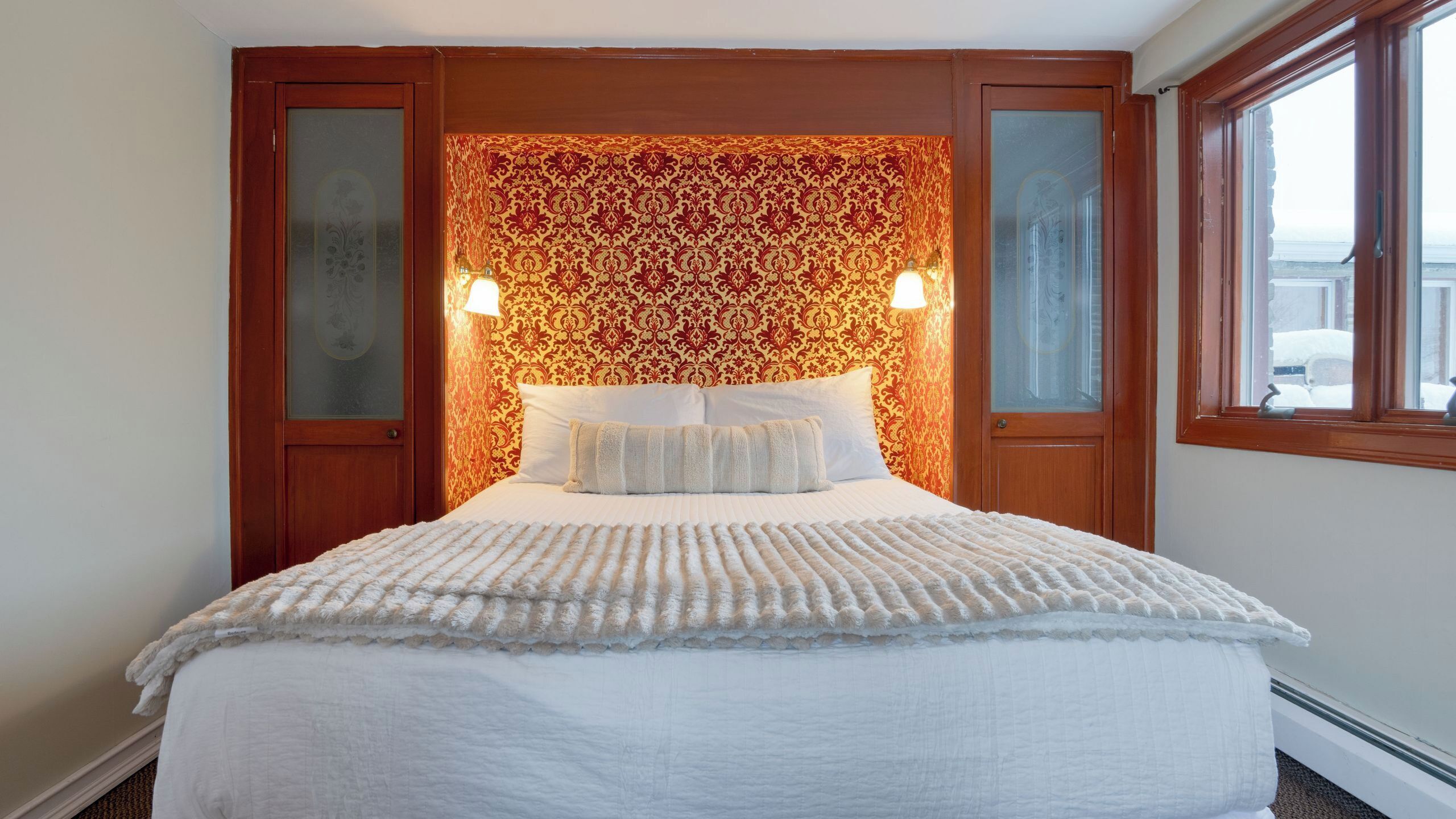 A hotel room with a large bed, two bedside lamps, a patterned wallpaper headboard, and a window with a view.