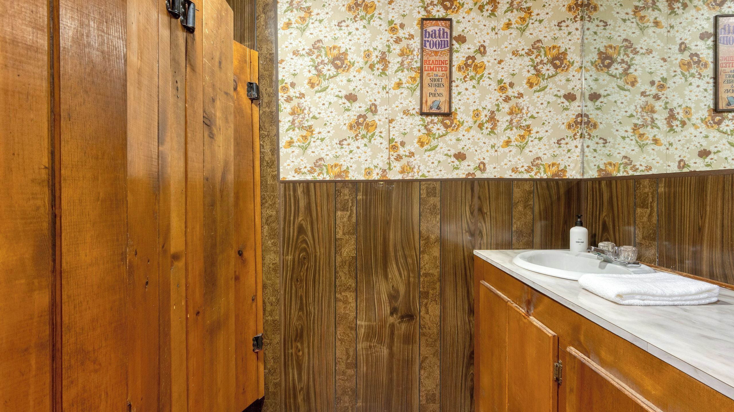 Une salle de bain avec des meubles en bois, un papier peint à motifs floraux et un lavabo doté d’un plan de travail blanc.