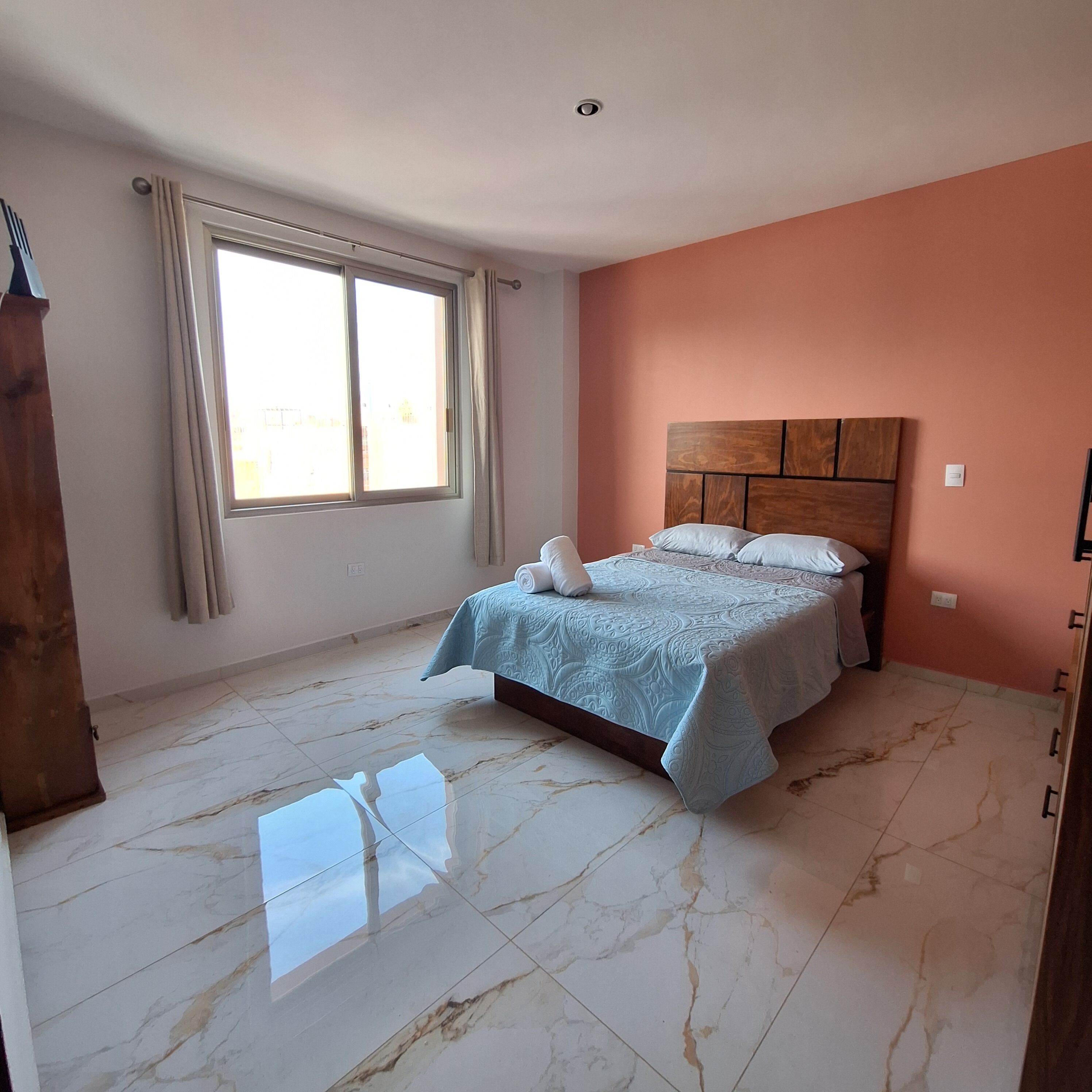 Une chambre à coucher avec un lit, une fenêtre avec des rideaux, une tête de lit en bois et un plancher en marbre.