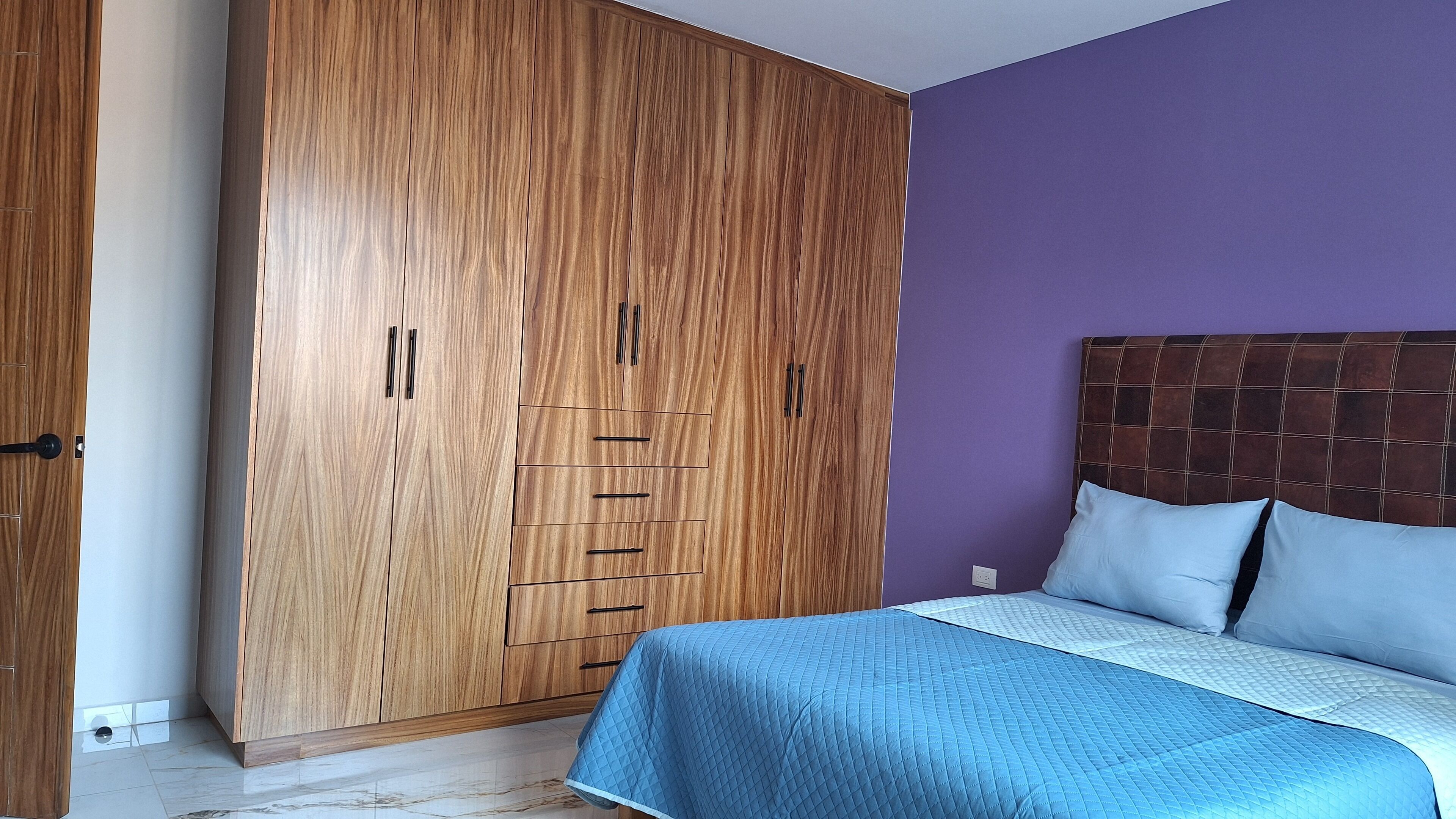 A bedroom with a large wardrobe, a bed with a blue quilt, and a purple wall.