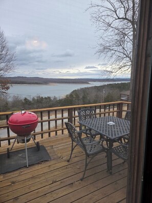 A deck with a grill and outdoor furniture overlooking a lake.