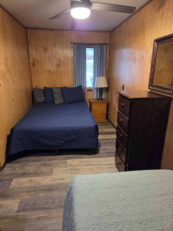 A bedroom with a bed, a dresser, a nightstand, a lamp, a ceiling fan, and a window with curtains.