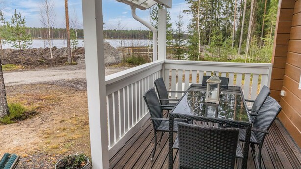 A covered outdoor deck with a glass-top table and black wicker chairs, overlooking a lake and forest. - Villa aleksandra by Interhome (Juva)