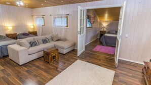Ein gemütliches Wohnzimmer mit Sofa, Couchtisch und Teppich. Zwei Fenster mit Vorhängen, eine Tür zu einem anderen Raum und eine Holzbalkendecke.