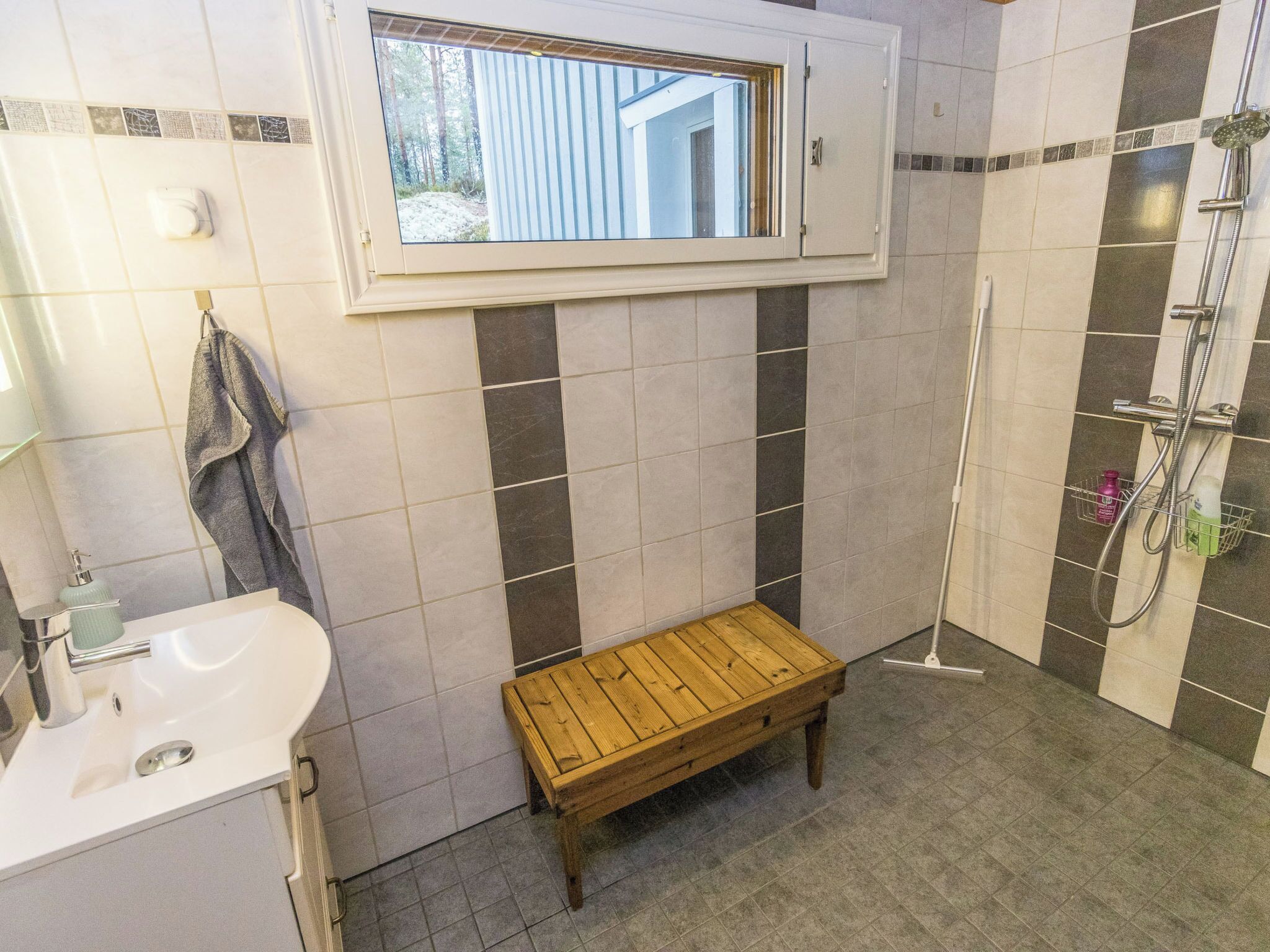 Une salle de bain avec un lavabo blanc, un banc en bois, une douche équipée d’un pommeau de douche à main et une fenêtre donnant sur un immeuble.