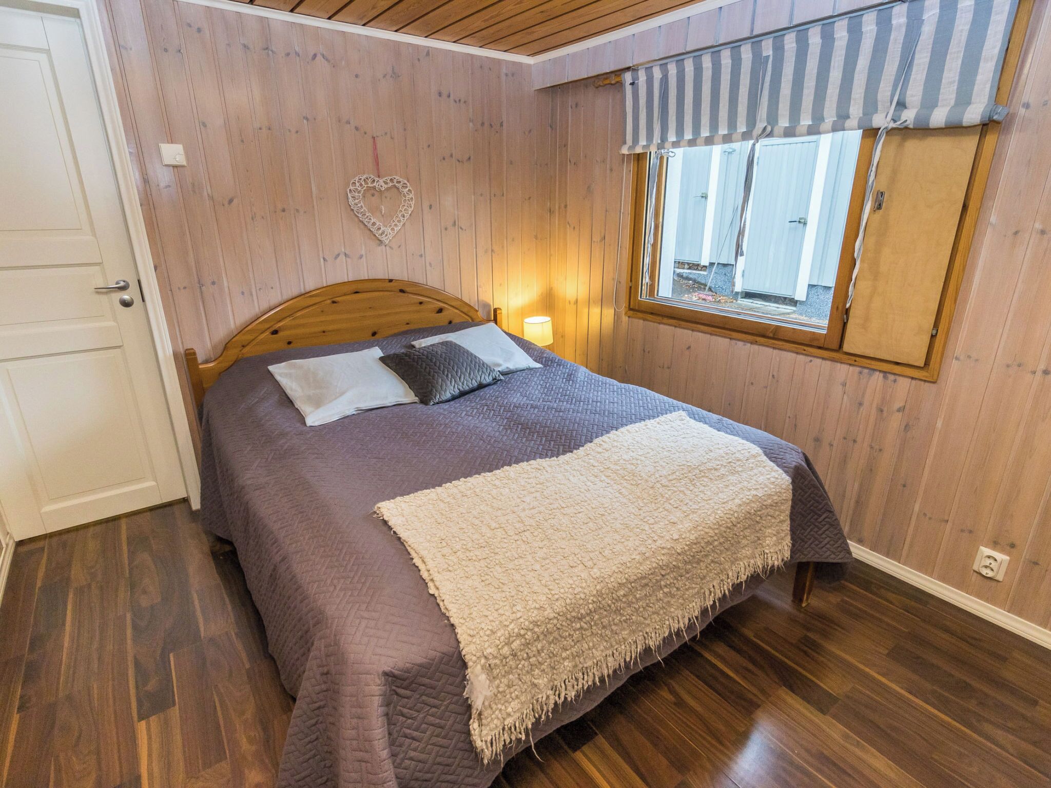 Un dormitorio con cabecera de madera, cama con colcha púrpura, manta beige, decoración con forma de corazón en la pared y ventana con cortina a rayas.