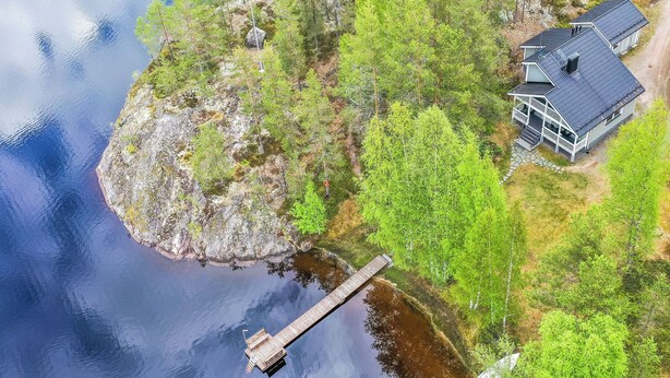 Ein Anwesen am Seeufer mit einer Anlegestelle, die ins Wasser ragt, umgeben von dichtem Grün und einem Haus mit dunklem Dach.
