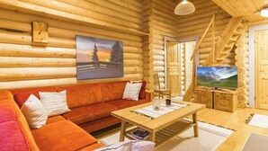 L’intérieur d’une cabane en rondins, avec un plancher en bois, un canapé, une table basse, un téléviseur et un tableau représentant un paysage.