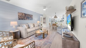A modern living room with a sofa, coffee table, and chairs, a dining area with a table and chairs, and a kitchen in the background. - Sunset Walks & Shelling Days at Seashells 9! Deeded beach access! (Sanibel)