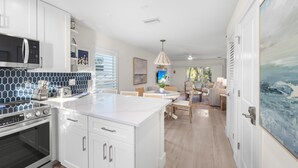 A modern kitchen with white cabinetry, a built-in oven, and a microwave. There is a dining area with a table and chairs, and a large painting on the wall. - Sunset Walks & Shelling Days at Seashells 9! Deeded beach access! (Sanibel)